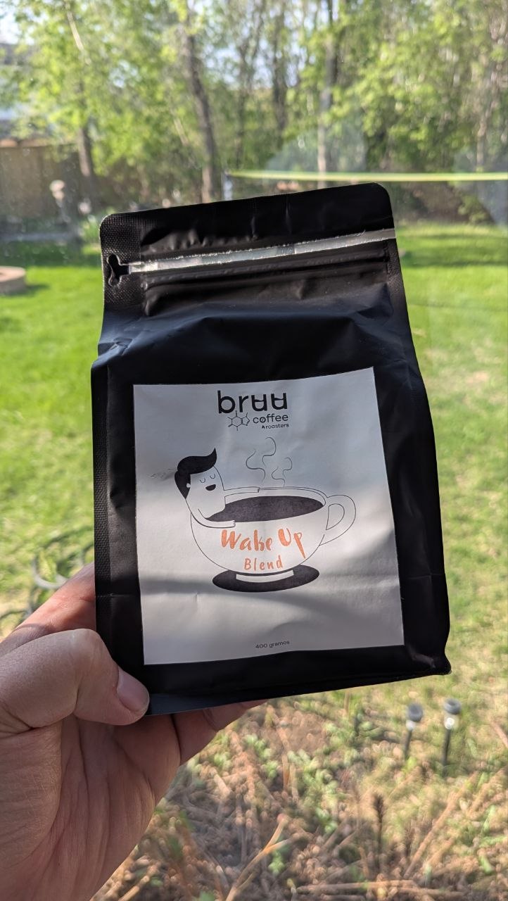 A black resealable coffee bag with a white label showing the "Wake Up Blend" branding by bruu coffee Roasters, held outdoors against a sunny green background