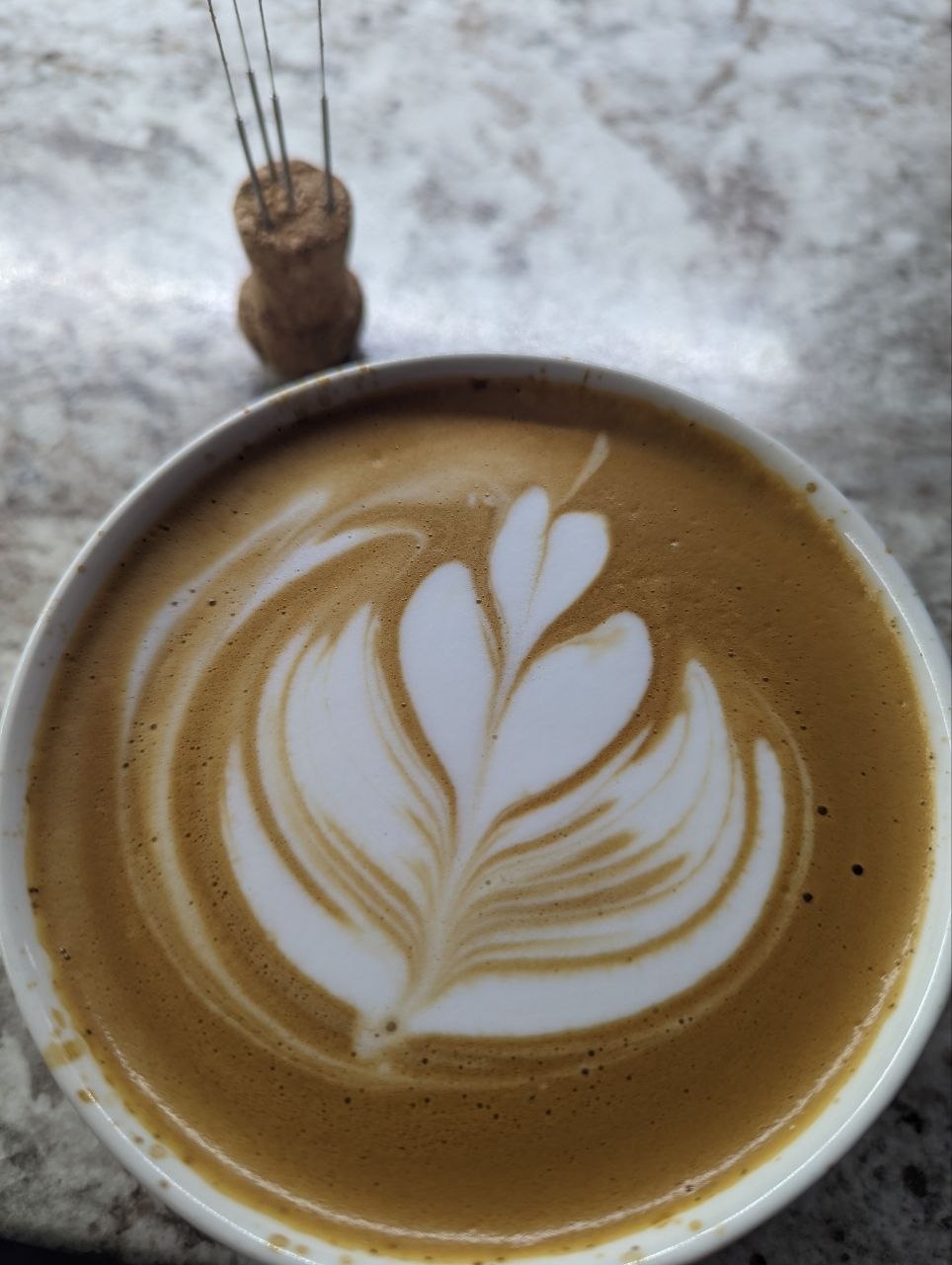 A cup of coffee with intricate latte art on a marbled surface, featuring a rosetta pattern and a small whisk in the background